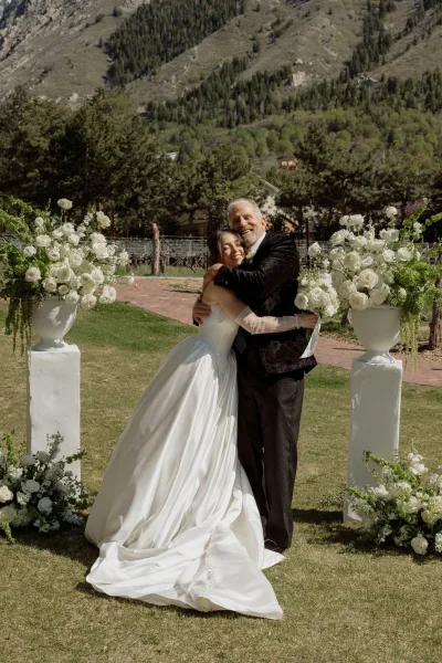 Bride and father portrait during a tender father-daughter hug, the bride in sheer gloves, with a mountain range and evergreens beyond.