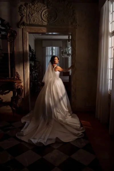 Bridal portrait of a bride in a looking-back pose, her button-back gown framed by an ornate doorway with carved molding and checkered floor.