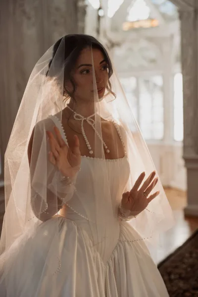 Bridal portrait of a bride under veil wearing a pearl choker necklace, posed by ornate windows with soft indoor sunlight.
