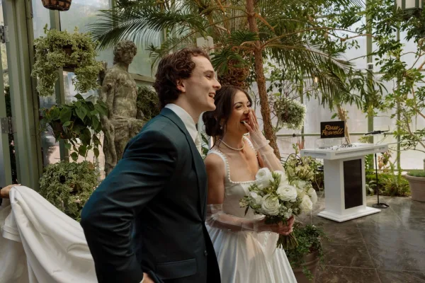 Couple portrait of bride and groom laughing as she holds a white and green bridal bouquet in a lush greenhouse framed by palm fronds and glass walls.