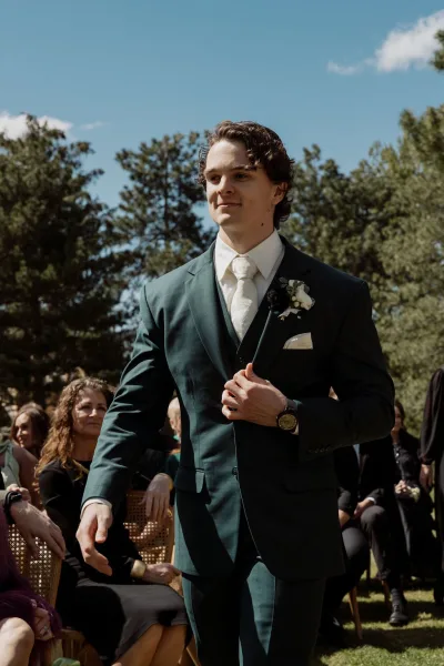 Ceremony processional in a candid ceremony moment as a person walks the aisle, boutonniere catching sun on lawn with seated guests.