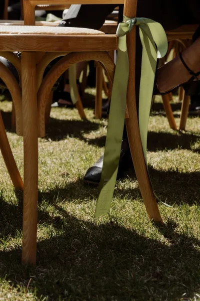 Ceremony chair decor with ribbon chair ties, a satin ribbon bow trailing from the chair back, on a grass lawn with seated guests beyond.