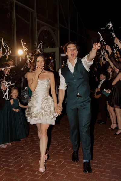 Wedding send off unfolds in a joyful ribbon wand exit as newlyweds walk hand in hand, the bride in a short bridal dress on a brick walkway at night.