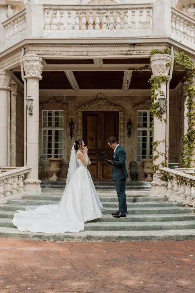 First look moment during a private vow exchange on stone steps by a mansion facade, as the groom reads from a vow book.