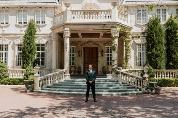 Groom portrait on stone mansion steps, standing in a tailored suit with hands clasped, columns and ivy framing grand wooden doors behind.