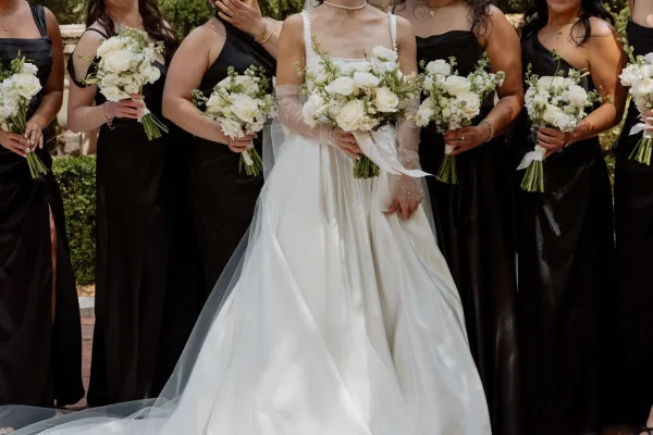 Bridal party portrait of the bride and bridesmaids holding white rose bouquets along a brick walkway framed by lush garden hedges.