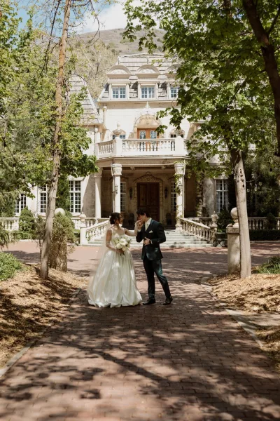 Couple portrait with the groom kissing bride's hand, her bridal bouquet held close, on stone steps before a grand mansion facade.