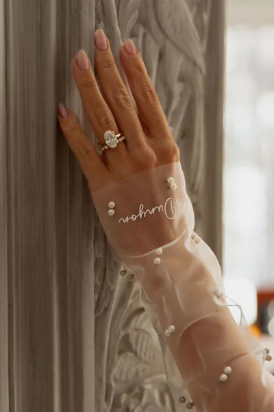 Engagement ring, an oval engagement ring gleaming on a pearl-embellished sheer glove, with carved stone column relief in soft focus behind.
