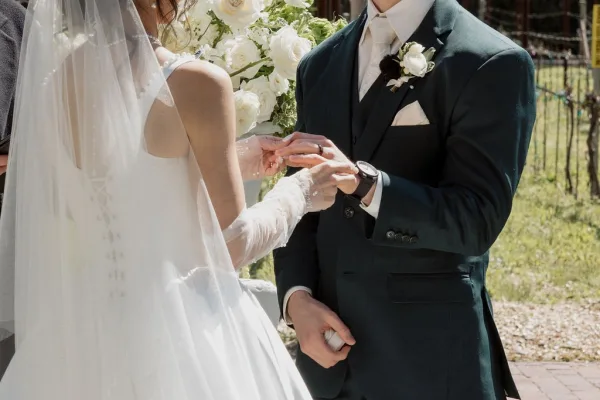 Wedding ring exchange during an outdoor ceremony moment, bride in sheer bridal gloves slides a band onto the groom’s hand amid garden greenery.