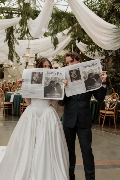 Couple portrait of a bride and groom with a custom newspaper, framed by draped ceilings, chandeliers, and hanging greenery at the reception.