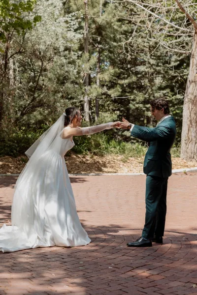 First look moment during an outdoor first look as the bride in sheer bridal gloves meets her groom on a brick path amid tall forest trees.