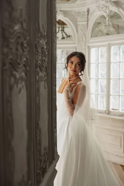 Bridal portrait of a bride with veil posed against ornate wall paneling and tall windows, with plaster details and wood floor in view.