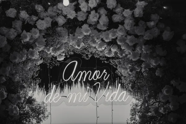 Wedding neon sign reading “amor” glowing on a dark wall, framed by rose garland and baby’s breath with hanging greenery above