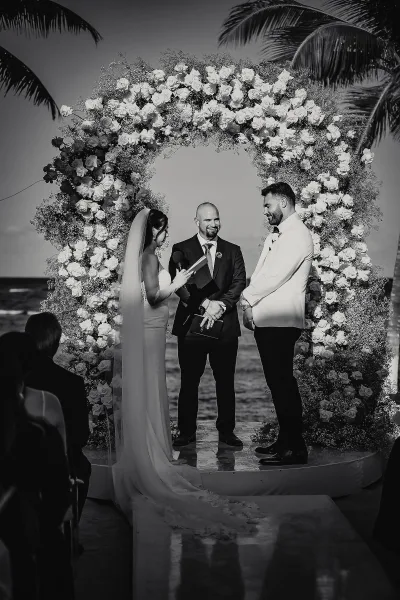 Wedding vows as bride reads into a microphone beside groom under a rose floral arch, long veil flowing, ocean and palms behind