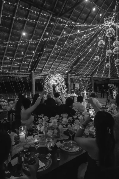 Wedding reception decor with string lights and disco balls overhead, floral arch and candlelit tables under a thatched ceiling with guests seated