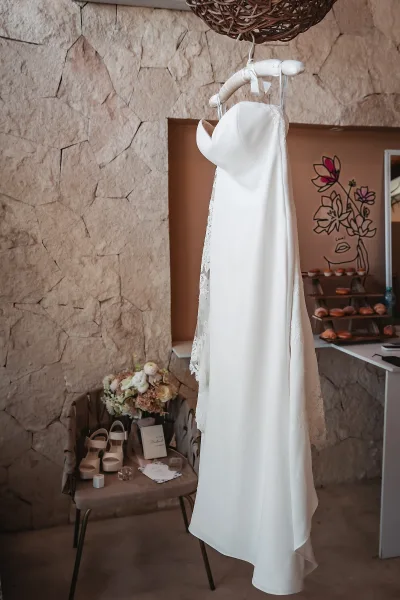Wedding dress on hanger with lace trim above bridal shoes, bouquet and invitations on a chair, set against a stone wall background