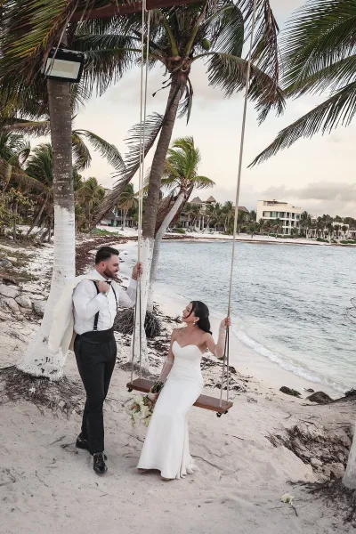 Couple portrait on a palm tree swing, bride in strapless wedding dress with bouquet and groom in suspenders on a sandy beach shoreline