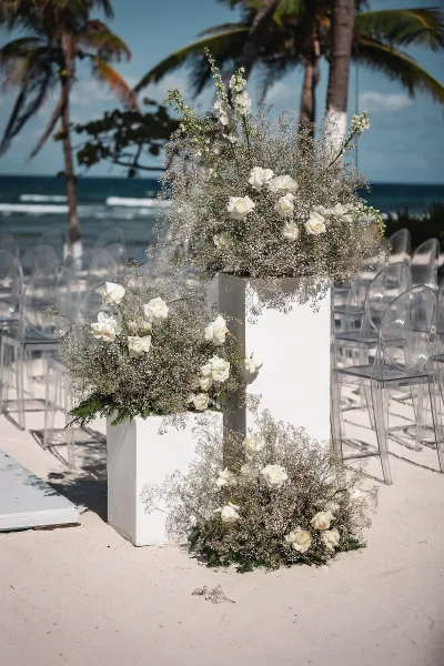 Ceremony floral arrangements of white rose blooms and baby's breath on tall pedestals and ground clusters beside clear chairs on a sandy beach
