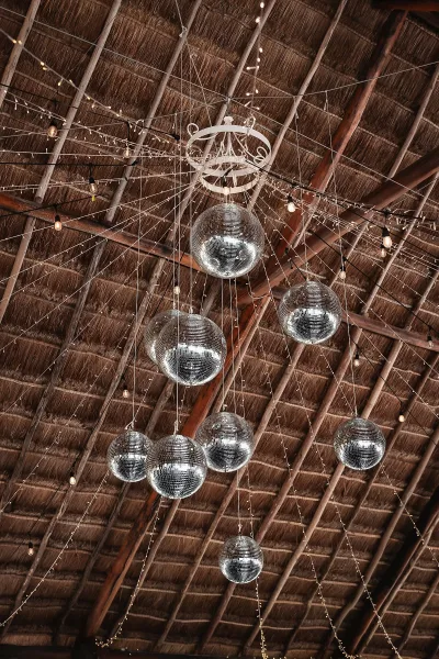 Reception lighting decor with disco ball wedding decor and warm string lights hanging from a thatched ceiling with wooden beams above the dance floor