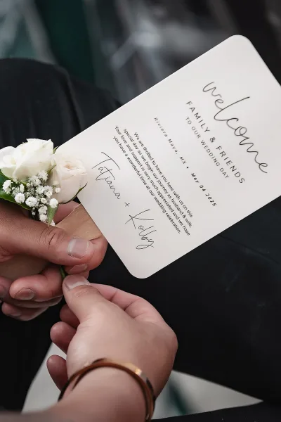 Wedding welcome card with calligraphy held by a groom in black suit, blush rose boutonniere with baby's breath against dark fabric
