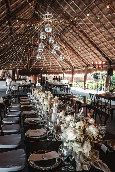 Reception tablescape on a long banquet table wedding with white floral centerpieces, candles, and string lights under a thatched roof pavilion