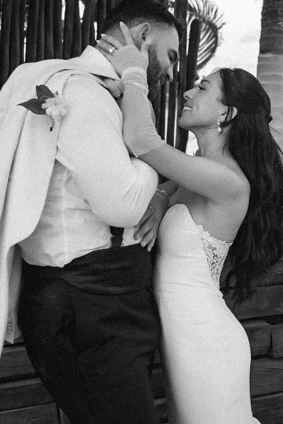 Wedding couple portrait with a tender forehead kiss as the bride holds the groom’s face, her lace dress and veil framed by palm leaves on a wood deck