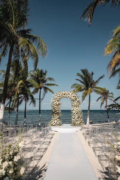 Ceremony setup for a beach wedding ceremony with a round white rose and greenery arch, clear chairs, and white aisle runner by the ocean