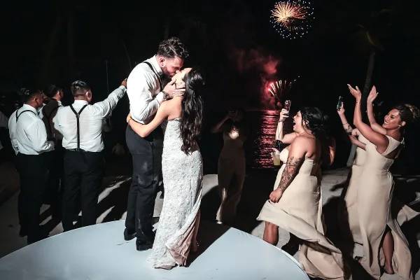 Wedding kiss as the couple dips on a beach at night beneath fireworks over the ocean, guests and bridesmaids cheering with phones raised