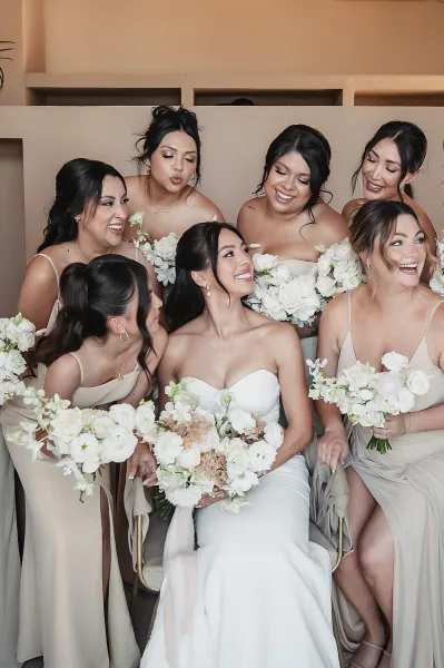 Bridesmaid group photo with bride in strapless gown, bridesmaids holding white rose bouquets, seated in a neutral room with shelves