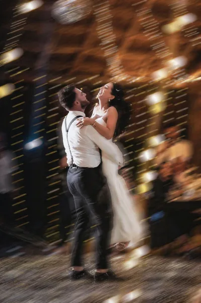First dance moment as groom in suspenders lifts bride in a wedding dress amid confetti, with cheering crowd under warm string lights