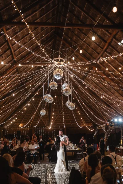 First dance under string lights as bride in a wedding dress and groom in tuxedo sway on the dance floor beneath disco balls in a rustic hall