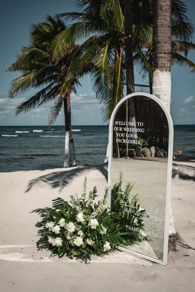 Wedding welcome sign on an arched mirror wedding welcome sign with white roses and greenery, set on a sandy beach with ocean and palm trees