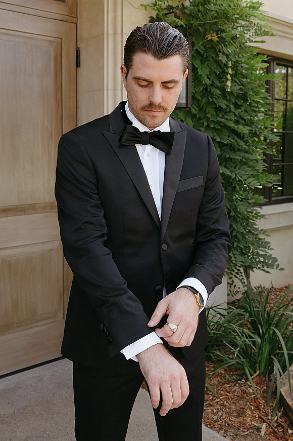 Groom portrait in a groom tuxedo, adjusting cufflinks with wristwatch and ring, standing by a stone building and wooden door outdoors