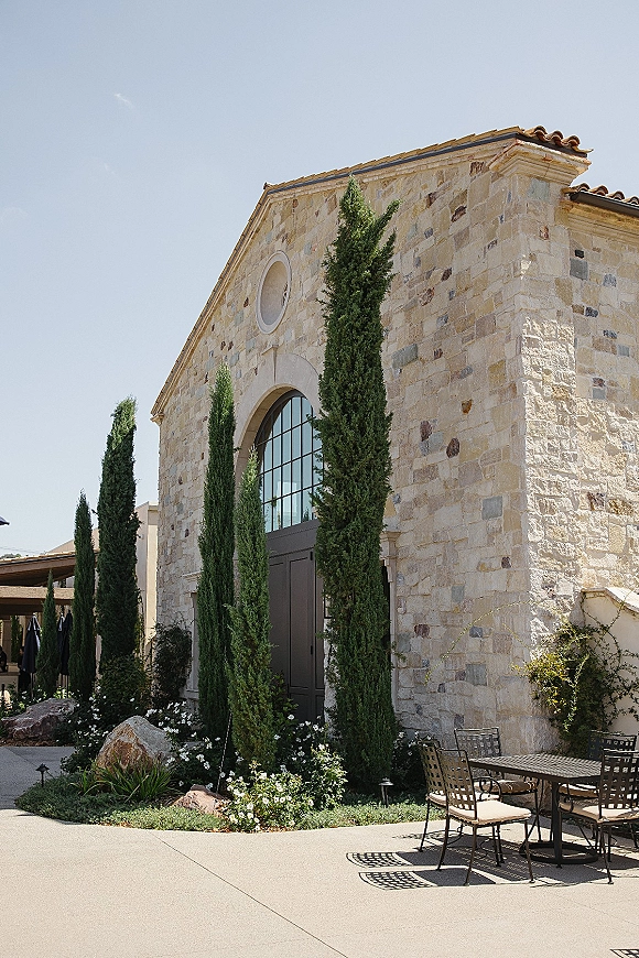 Wedding venue exterior with stone wedding venue facade, arched window and double doors, cypress-lined courtyard walkway and patio umbrellas under blue sky