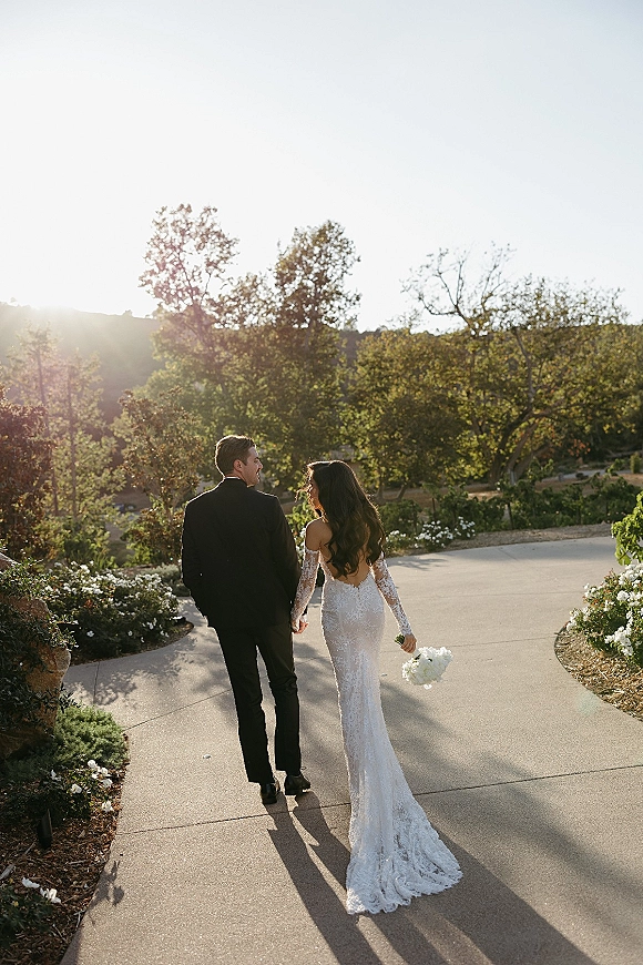 Couple portrait of bride and groom walking away hand in hand on a sunlit garden path, bride in long sleeve lace gown holding bouquet