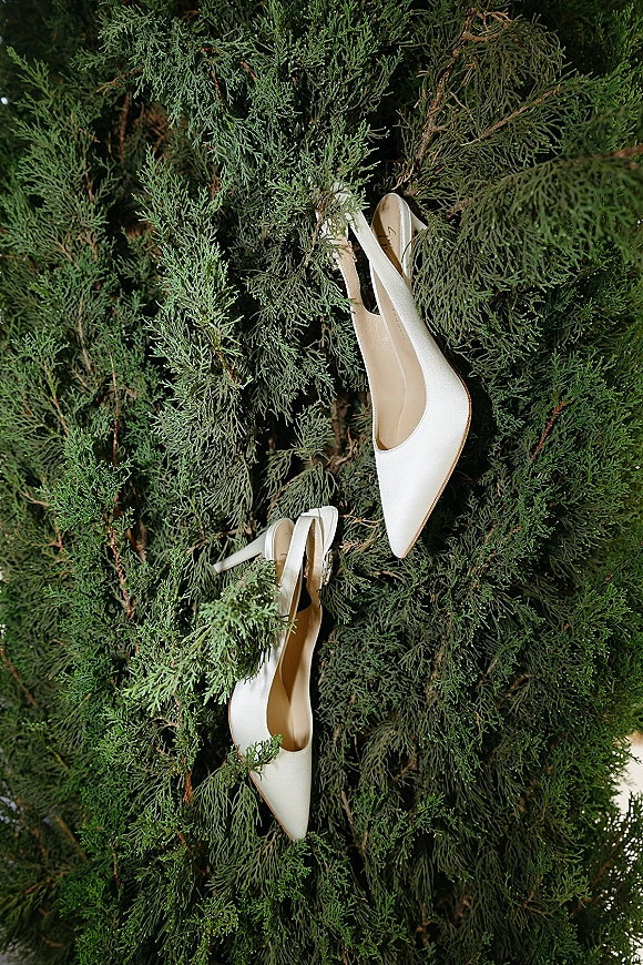 Bridal shoes arranged as a wedding shoes flatlay, white pointed-toe heels resting on lush evergreen branches and greenery foliage