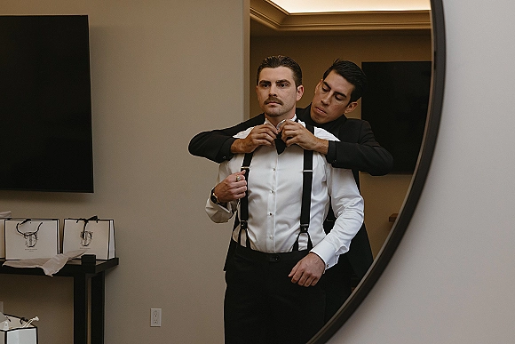 Groom getting ready as a groomsman adjusts his black bow tie, suspenders and wristwatch visible in a hotel room mirror setting
