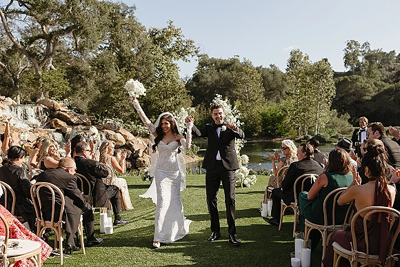 Wedding recessional as bride and groom walk the aisle, bride raising bouquet overhead by pond and rock waterfall under blue sky