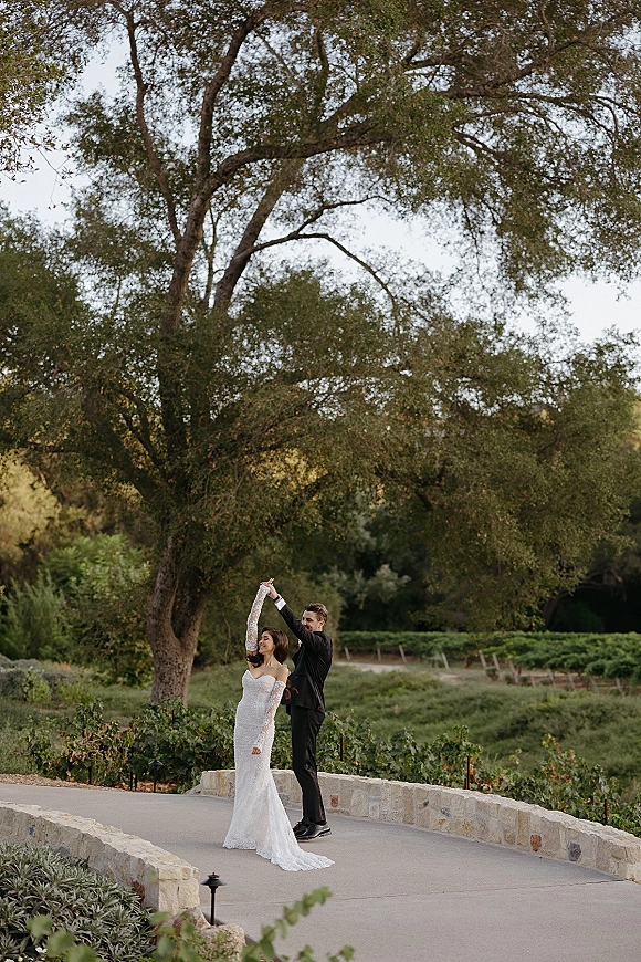 Couple portrait of bride and groom twirl on a paved vineyard path, her lace sleeve gown and dress train flowing beside his black tuxedo