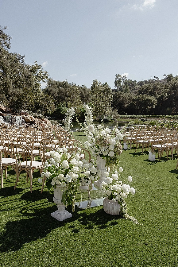 Ceremony setup with outdoor ceremony seating, clear aisle runner lined with white florals and candles on a lawn by a pond and waterfall