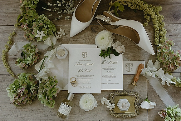Invitation suite wedding invitation flat lay with calligraphy cards, wax seal, silk ribbon, rings, perfume, and heels on a wood tabletop