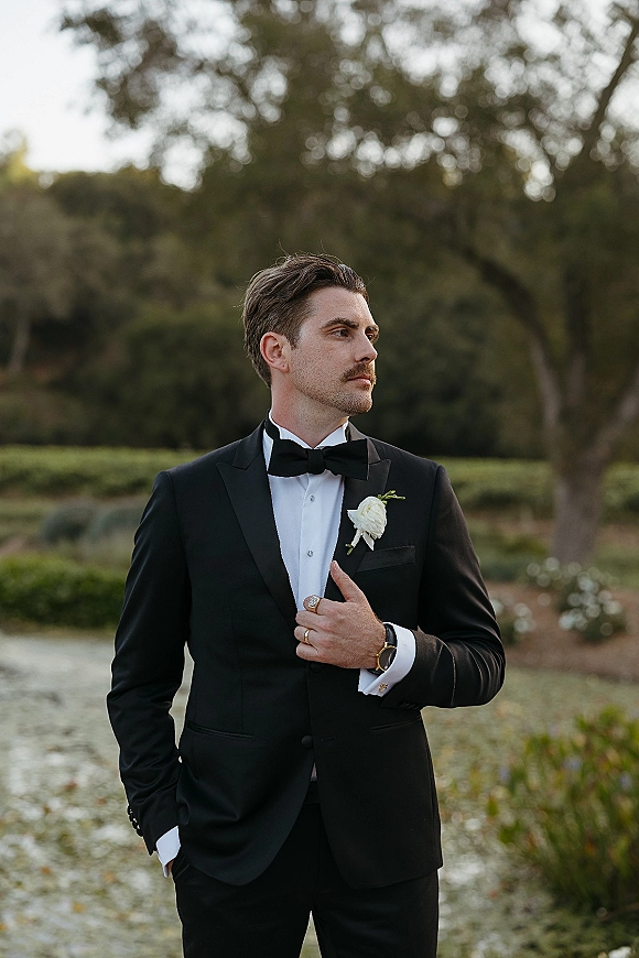 Groom portrait in a black tuxedo groom look with bow tie and white rose boutonniere, standing in a blurred garden backdrop
