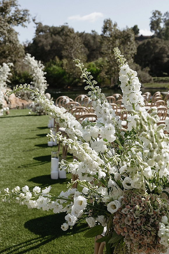 Ceremony aisle decor with outdoor wedding aisle lined by white hydrangea florals and glass candle holders on a lawn by a pond
