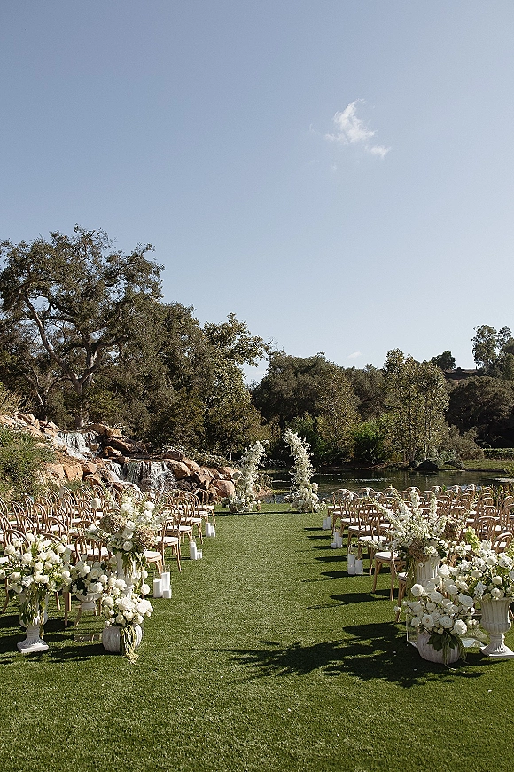Ceremony setup with white floral arrangements lining an aisle runner and curved back chairs beside a pond and waterfall backdrop