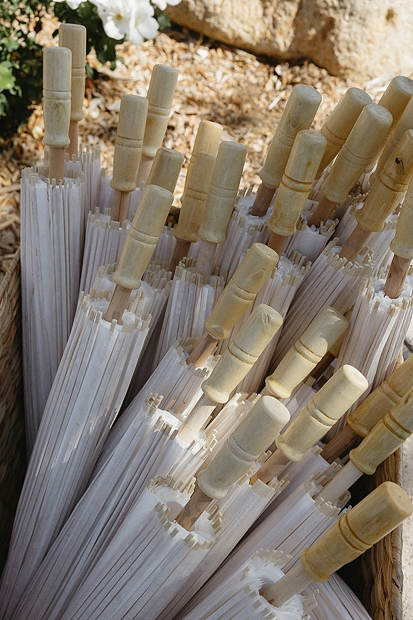 Wedding parasols with white paper canopies and wooden handles arranged in a wicker basket on mulch near stone and greenery outdoors