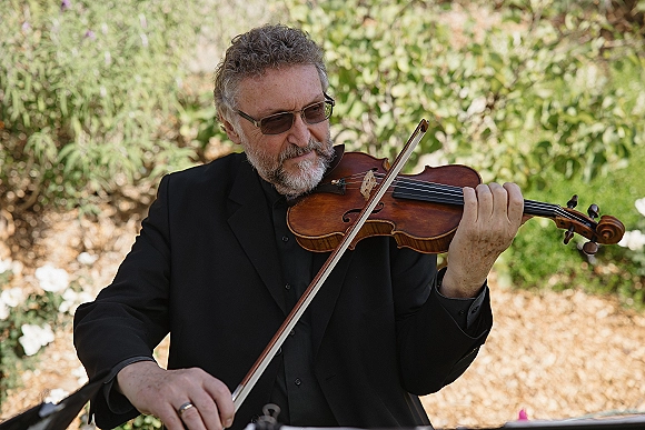 Wedding violinist in a black suit and sunglasses playing with bow and wedding band ring visible against lush garden greenery backdrop