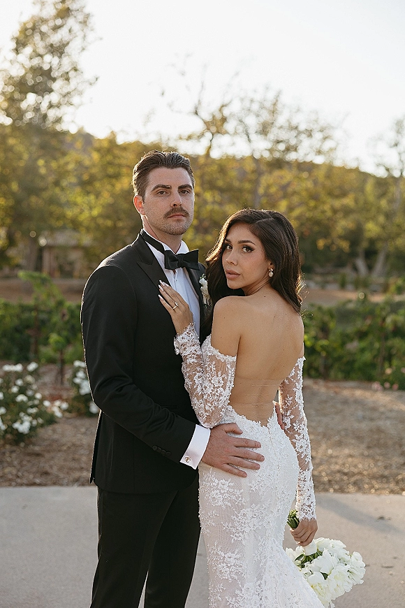 Couple portrait of bride and groom pose, her hand on his chest, holding a white bouquet on a garden path at sunset
