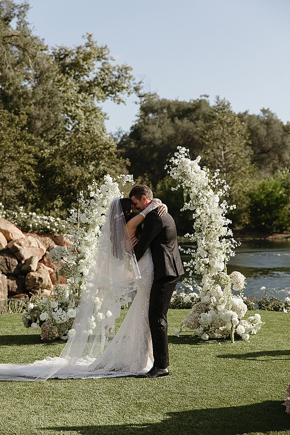 Wedding kiss under a white floral arch with veil draped over the couple, lakeside lawn and trees behind in natural light
