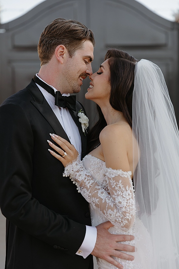Wedding couple portrait of bride and groom close up touching foreheads, her long veil and lace off-shoulder gown against a neutral wall backdrop