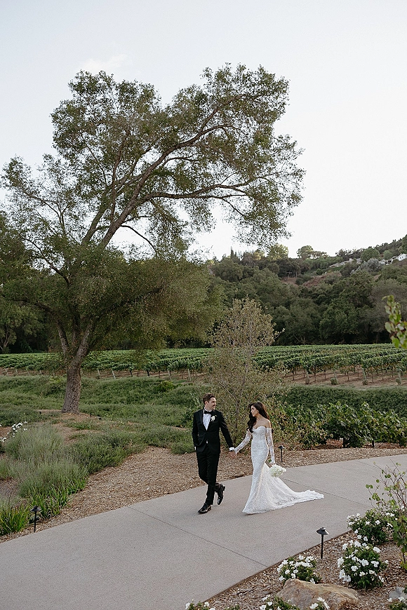 Couple portrait of bride and groom walking hand in hand, her veil and bouquet flowing, along a vineyard path with hills behind
